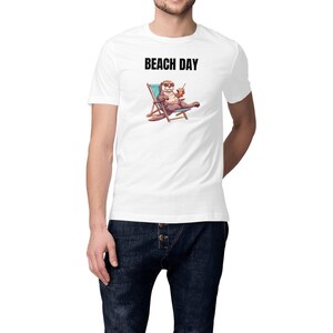 Peut inclure: Un t-shirt blanc à manches courtes avec un motif représentant une loutre portant des lunettes de soleil, allongée sur une chaise de plage bleue en tenant une boisson. Au-dessus, le texte BEACH DAY est imprimé en lettres capitales noires.