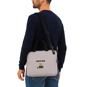 Peut inclure: Une sacoche pour ordinateur portable gris clair avec des poignées noires et une bandoulière réglable. L'avant présente un graphique d'un chat orange portant des lunettes, assis devant un ordinateur avec une tasse, et le texte CAMPUS MODE.