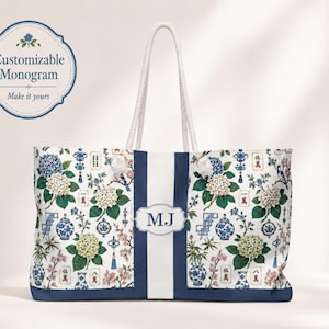 May include: A white tote bag with rope handles featuring a blue and white chinoiserie pattern of hydrangea flowers, vases, and mahjong tiles. A central vertical navy blue stripe displays a white label with the monogram MJ in navy blue letters.