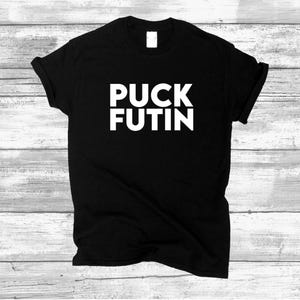 May include: Black t-shirt with the white text "PUCK FUTIN" in a bold, sans-serif font. The shirt is displayed on a light-colored, wood-grain background. The short-sleeved shirt is folded and has a tag.