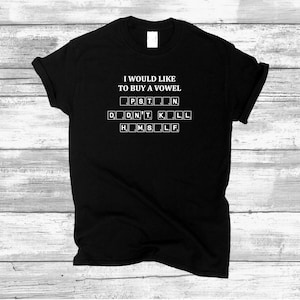 Może przedstawiać: Czarna koszulka z białym napisem. Tekst brzmi "I WOULD LIKE TO BUY A VOWEL" nad serią płytek z literami, które wymawiają "PST N, DONT K LL, H MS LF". Koszulka jest prezentowana na jasnej powierzchni z usłojeniem drewna.