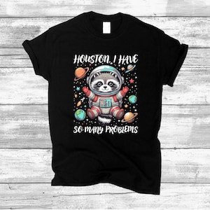 May include: Black t-shirt featuring a cartoon raccoon dressed as an astronaut, surrounded by planets. The text "Houston, I have so many problems" is printed on the shirt.