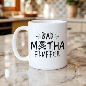 May include: White ceramic mug with a black graphic that reads "BAD MOTHA FLUFFEr" with a skull and crossbones. The mug has a curved handle and is sitting on a marble countertop.