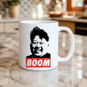Può includere: Tazza in ceramica bianca con una grafica in bianco e nero di una persona sorridente. La parola "BOOM" è stampata in un rettangolo rosso sotto la grafica. La tazza ha un manico curvo.