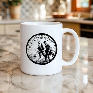 Può includere: Tazza in ceramica bianca con un'illustrazione in bianco e nero di due persone che usano un metal detector, circondata dalla parola "DETELTORISTS". La tazza ha un manico curvo. Lo sfondo è sfocato.