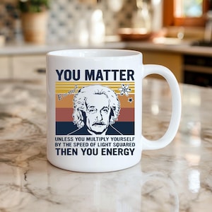 May include: White ceramic mug with a graphic of Albert Einstein and the text "YOU MATTER" and a physics equation. The mug also includes the text "UNLESS YOU MULTIPLY YOURSELF BY THE SPEED OF LIGHT SQUARED THEN YOU ENERGY."