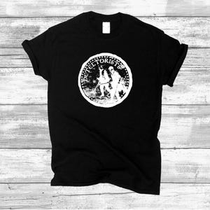Può includere: T-shirt nero con grafica bianca. La grafica è un cerchio con la scritta "DETELTORISTS" sul bordo e un'illustrazione dettagliata al centro. La maglietta ha maniche corte e girocollo.