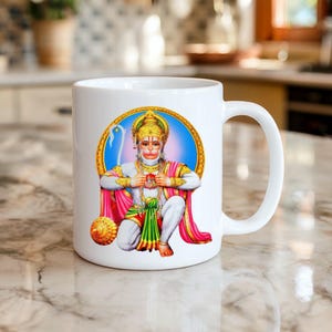 May include: White ceramic mug featuring a colorful illustration of a Hindu deity, Hanuman. The artwork includes gold, red, and blue accents, set against a blue background. The mug has a curved handle and is suitable for hot beverages.
