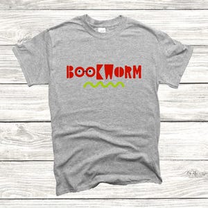 Op de afbeelding: Een lichtgrijs t-shirt met het woord "BOOKWORM" in rode letters en een groen zigzagontwerp eronder. Het woord "BOOKWORM" heeft een speels ontwerp, waarbij de "O"'s zijn vervangen door ogen.