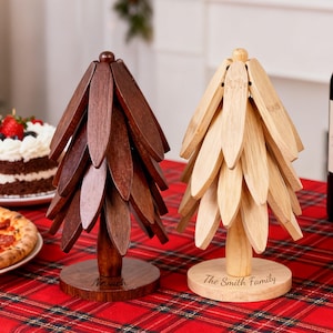 Peut inclure: Deux décorations d'arbre de Noël en bois, une marron foncé et une marron clair, sont posées sur une nappe à carreaux rouges. L'arbre marron foncé porte le nom "Noah" gravé sur sa base, tandis que l'arbre marron clair porte "The Smith Family".