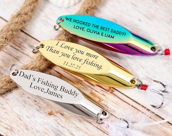 May include: Three fishing lures with personalized text. One lure is silver with "Dad's Fishing Buddy Love, James". Another is gold with "I Love you more Than you love fishing. 11.27.25". The third is teal with "WE HOOKED THE BEST DADDY! LOVE, OLIVIA & LIAM".