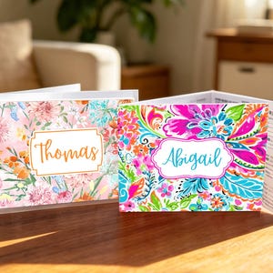 May include: Two folded cards with floral designs. One card has a pink and orange floral pattern with the name "Thomas" in an orange frame. The other card has a vibrant floral pattern with the name "Abigail" in a blue frame.