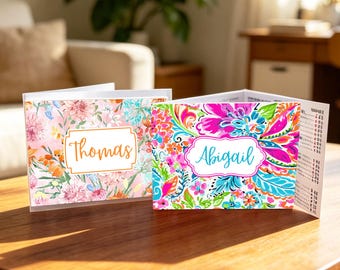 May include: Two folded cards with floral designs. One card has a pink and orange floral pattern with the name "Thomas" in an orange frame. The other card has a vibrant floral pattern with the name "Abigail" in a blue frame.