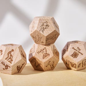 Puede incluir: Cuatro dados dodecaédricos de madera con ilustraciones y texto grabados. Los dados presentan imágenes de personas, comida y las palabras "Play Board Games" e "Italian". Los dados están apilados sobre una superficie de madera clara.