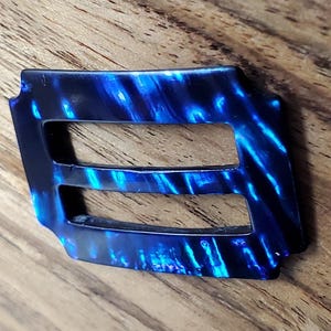 May include: A rectangular belt buckle featuring a glossy, marbled pattern in shades of deep navy blue and bright electric blue. The buckle has two horizontal slots for threading a belt strap, with a smooth, polished finish and defined edges.
