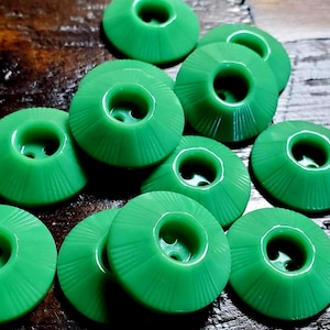 May include: A collection of bright green circular buttons, each featuring a raised center with two holes and a series of fine, radiating lines etched into the sloped surface, creating a sunburst pattern on the face of each individual button.