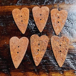 May include: Six heart-shaped buttons with a textured, rusted brown finish. Each button features two vertical holes in the center for attachment. The buttons are arranged in two rows of three, displaying a weathered, metallic appearance.