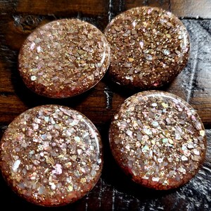 May include: Four circular buttons featuring a textured, rose gold glitter finish with embedded iridescent flecks. Each button has a smooth, rounded surface and a reflective, sparkling appearance that catches the light, creating a multi-dimensional effect.