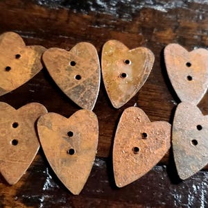 May include: Eight heart-shaped buttons with a weathered, rusted copper-toned finish. Each button features two small holes in the center for attachment. The surface of each button displays a mottled, textured appearance with varying shades of brown.