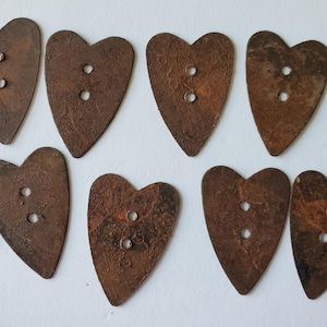 May include: A set of seven heart-shaped buttons with a dark brown, textured, oxidized metal finish. Each button features two circular holes positioned vertically in the center of the heart shape, displaying a weathered and rustic appearance.