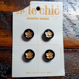 May include: A set of four round black buttons on a white Le Chic display card. Each button features a raised gold-colored maple leaf design. The card includes the text Le Chic, Guaranteed Washable, 5679-22, 9/16 inch, and Western Germany.