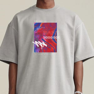 Op de afbeelding: Een grijs t-shirt met een kleurrijke abstracte grafische afbeelding op de voorkant. De afbeelding bevat rode, blauwe en witte elementen met het woord "RAD" en andere tekst. Het shirt heeft korte mouwen en een relaxte pasvorm.
