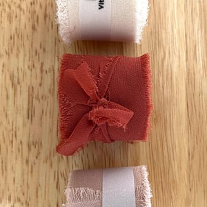 May include: Three rolls of fabric ribbon in cream, rust, and blush pink, each with frayed edges. The cream and blush pink rolls have a white label with the Vitalizart logo. The rust ribbon is tied in a bow.
