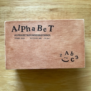 May include: A wooden stamp set labeled "Alpha Be T" with the text "ALPHABET & NUMBER & SYMBOL". The stamp size is 9x12x40 mm. The set includes 70 pieces. A small smiley face is next to the letters and numbers.