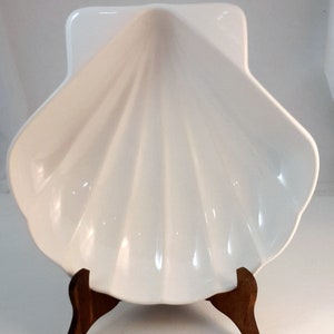 Puede incluir: Plato de servir de cerámica blanca con forma de concha marina con un soporte de madera. El plato está diseñado para parecerse a una gran concha de almeja.