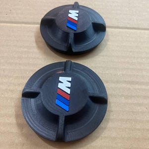 May include: Two black, circular ashtrays with the BMW M logo in the centre. The logo features the letter "M" in white, with red, blue, and light blue stripes. Each ashtray has three small grooves for resting cigarettes.