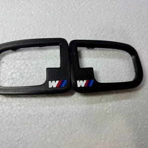 Może przedstawiać: Dwa czarne akcesoria samochodowe o prostokątnym kształcie i zakrzywionej krawędzi. Każdy ma białe logo "W" z czerwonymi i niebieskimi paskami. Prawdopodobnie części pojazdu.