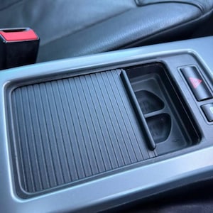 Puede incluir: Primer plano de la consola central de un coche, con una cubierta corredera negra y acanalada y dos portavasos. La consola está rodeada de un embellecedor gris claro y asientos de cuero negro. Se ve un botón rojo de luces de emergencia.