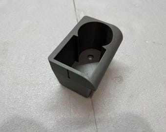 May include: A gray, 3D-printed cup holder with a rectangular base and a circular opening for a cup. The holder has a curved edge and a small hole in the bottom. The design is simple and functional.