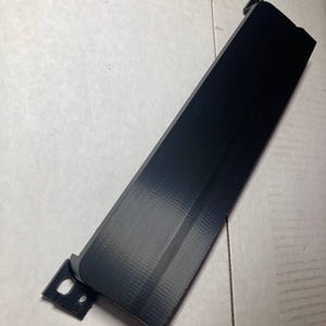May include: A black, rectangular plastic component with a textured surface. The component has a slightly angled design and a small, attached section with two holes. The item is likely a part of a larger product.