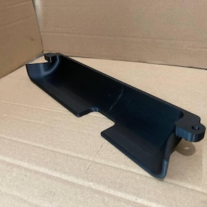 Peut inclure: Un composant en plastique noir, allongé avec un canal incurvé et une base plate. La pièce a une forme rectangulaire avec un bord arrondi et un petit trou circulaire à une extrémité. L'article fait probablement partie d'un assemblage plus grand.