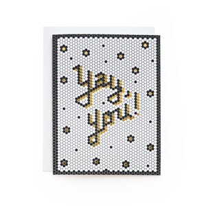 May include: A white card with a black border and a gold mosaic pattern. The words "yay you" are written in gold letters on the card.