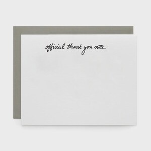 May include: A white card with the handwritten text "official thank you note" in black script. The card is set against a gray background, suggesting a stationery or greeting card product.