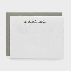 May include: A white stationery card with the handwritten phrase "a little note" in black script. The card is set against a gray envelope, creating a simple, minimalist design. The text is centered, offering space for a personal message.
