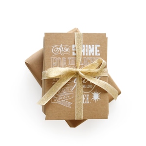 May include: Two brown paper wrapped gifts tied with a gold ribbon. The top gift has white text that reads "Arise, Shine for the Light is Upon You." The bottom gift has white text that reads "And the Lord is with thee."