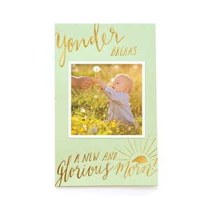 May include: A green card with gold lettering that says "Yonder Breaks a New and Glorious Morn!"  A photo of a baby reaching for a dandelion is in the center of the card.