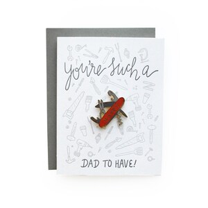 May include: A white card with a gray envelope. The card has a black and white illustration of tools and the text "you're such a dad to have!" A red and silver pocket knife pin is on the card.