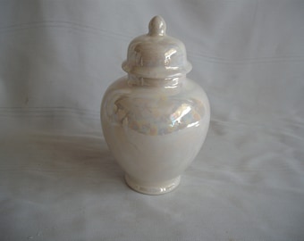 May include: A pearlescent white ceramic jar with a lid. The jar has a rounded body and a small, rounded lid with a knob on top. The surface has an iridescent sheen, reflecting colors. The jar is likely used for storage or decoration.