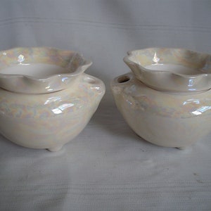 May include: Two off-white ceramic candle holders with an iridescent pearlescent sheen. Each has a round base with three small feet, a bowl-shaped body, and a smaller, scalloped-edge dish on top.