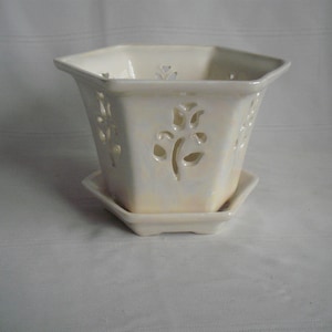 May include: A hexagonal off-white ceramic planter with an iridescent glaze finish. The planter features cut-out floral motifs on each side and sits on a matching hexagonal saucer. The design includes a wide rim and a tapered base structure.