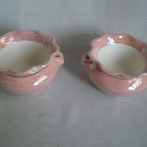 May include: Two matching pink ceramic bowls with a ruffled rim and small handles. The bowls have a pearlescent finish and a white interior. These decorative bowls are ideal for serving or display.