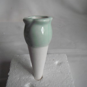 Könnte beinhalten: Eine Keramik-Blumenvase in Form einer Eiswaffel. Der obere Teil ist in einem glänzenden, blassen Minzgrün mit gewelltem Rand glasiert, während der untere Teil eine matte, unglasierte weiße Waffel mit einer strukturierten, porösen Oberfläche ist.