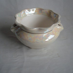 May include: A two-piece, off-white ceramic bowl set. The top bowl has a scalloped rim and iridescent accents. The bottom bowl has two small handles. The set is designed for decorative use.
