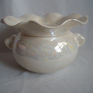 Op de afbeelding: Een parelmoerwitte keramische kom met een gerimpelde rand en twee kleine handvatten. De kom heeft een ronde basis en een deksel dat er goed op past. Het oppervlak heeft een subtiele iriserende glans.