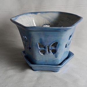Peut inclure: Un pot de fleurs en céramique bleu clair avec des découpes en forme de papillon. Le pot a une base carrée et une soucoupe assortie. Le pot mesure environ 15 cm de large et 13 cm de haut.