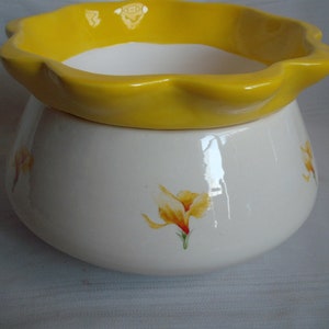 May include: A two-piece ceramic container. The base is white with yellow floral accents. The top is a bright yellow, scalloped rim. The container is likely for decorative or functional use, such as a planter or storage.